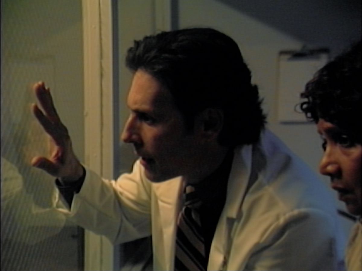 A man in a white lab coat presses a hand against an observation window and gapes as he looks into the next room in the V/H/S/85 segment Total Copy