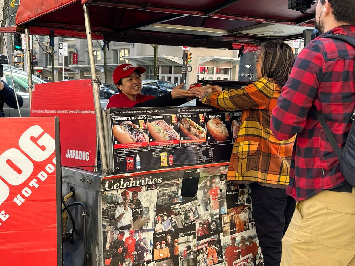 A vendor hands a hotdog to customers over the front of a food cart advertising various dishes.