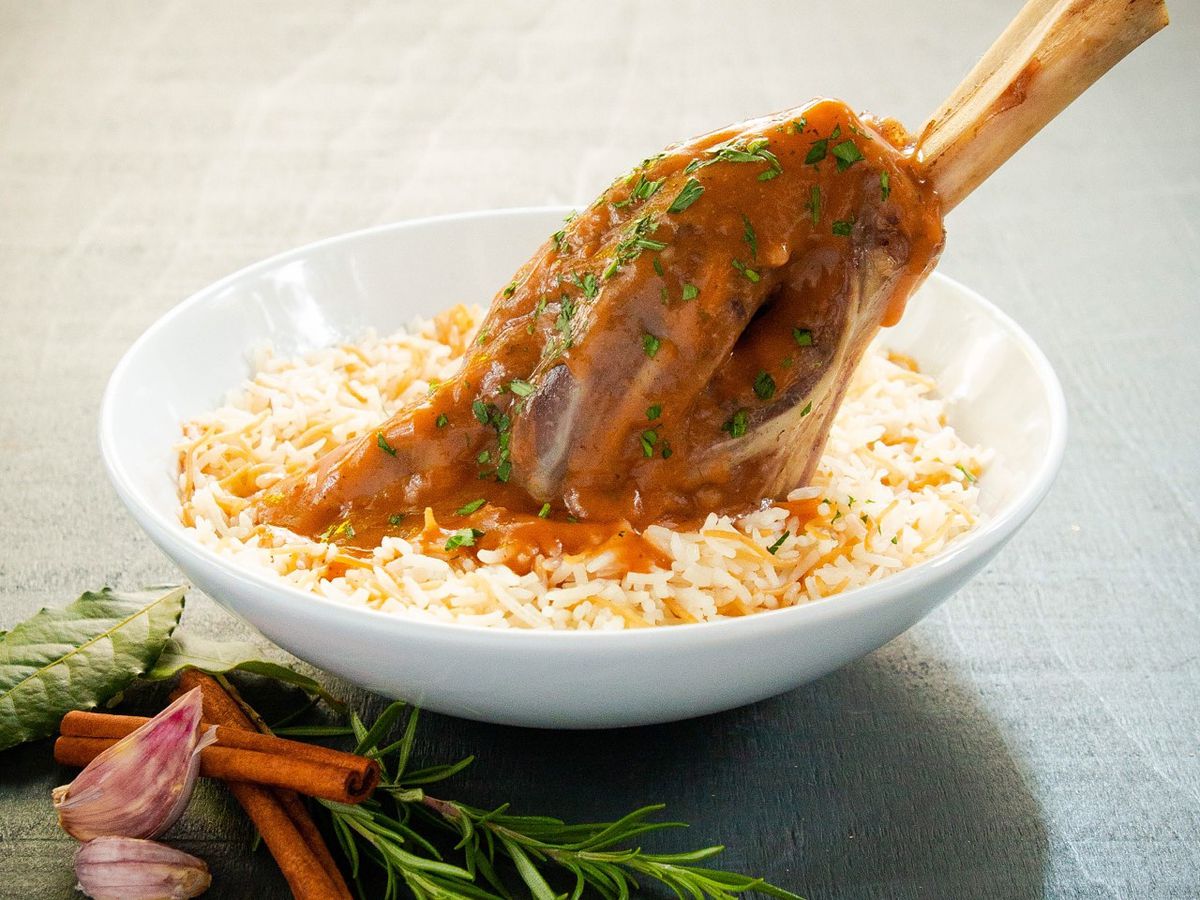 A lamb shank sits in a bowl of rice on a table with vegetables in the foreground. 