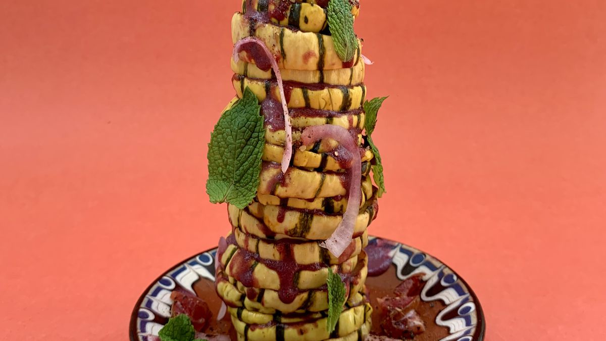 A tall stack of sliced squash garnished with mint leaves and red onion on a ceramic plate against a red backdrop.