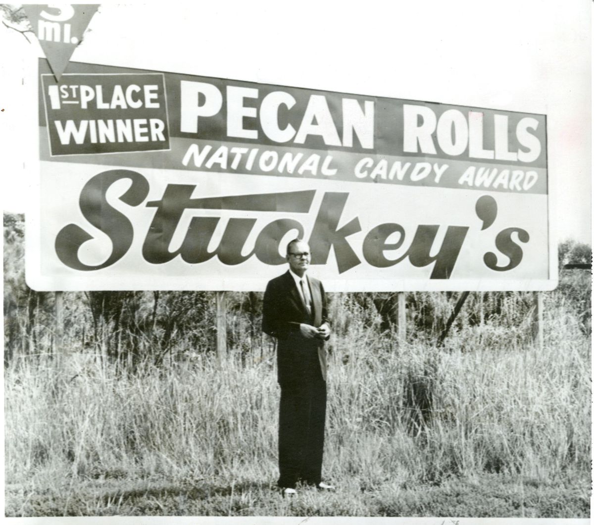 Black and white photo of W.S. “Sylvester”&nbsp;Stuckey, Sr. standing in front of a roadside billboard which reads, “First place winner. Pecan Rolls. National Candy Award. Stuckey’s.”