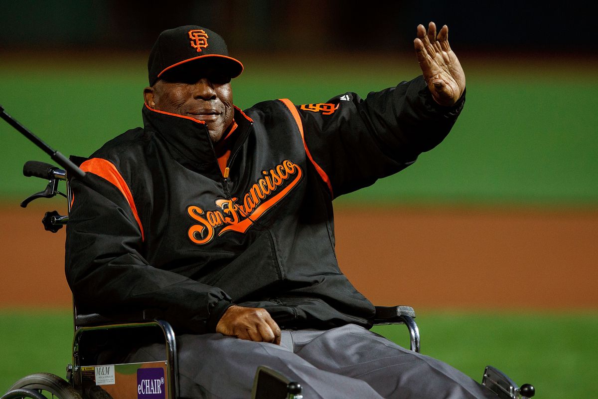Willie McCovey waiving to fans while in a wheelchair on the Giants field.