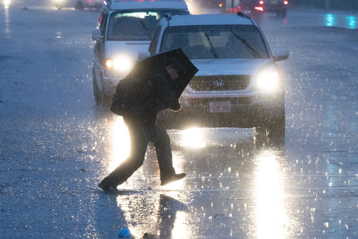 A person carrying an umbrella hunches underneath it as they cross the street under what looks like heavy rain. Cars with their windshield wipers and headlights turned on wait in the background.