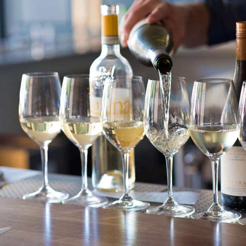 A hand behind the bar at Alma is pouring white wine into a line up of glasses
