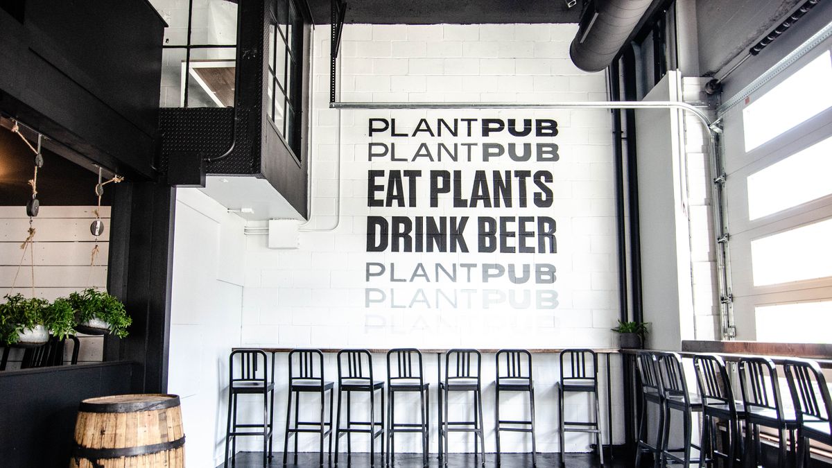 A casual, sun-lit restaurant features black lettering on a white wall that reads Plantpub Eat Plants Drink Beer. Plants and a decorative barrel are in the space.