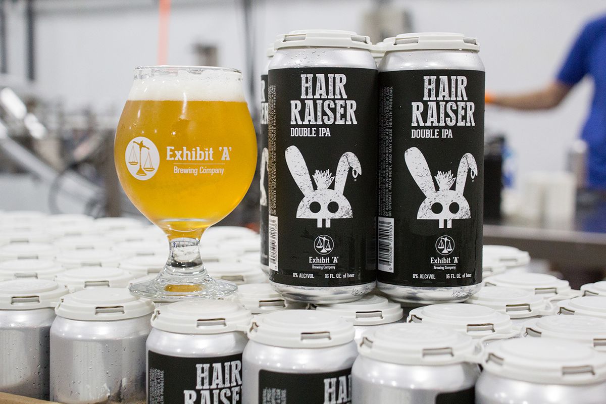 A full tulip glass sits alongside cans of Exhibit ‘A’ Brewing Company’s Hair Raiser Double IPA