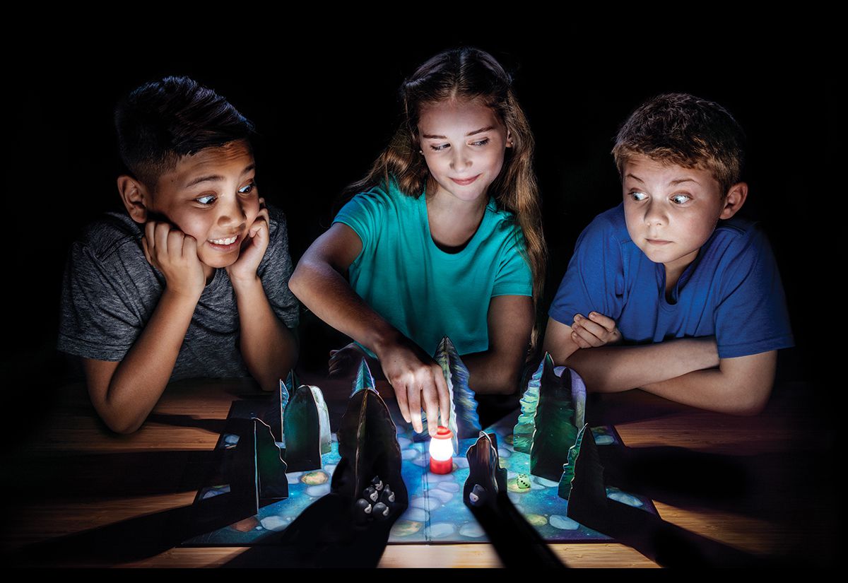 Children sit around a table in a dark room moving game pieces on the table. The pawn is shaped like a camping lantern, and cardboard trees cast shadows all around.