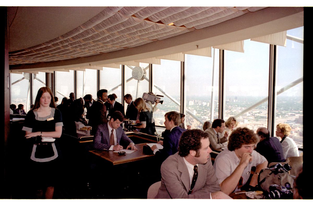 Inside a restaurant with an observation deck in Dallas’s Reunion Tower in 1978. Men and women in garb from the era sit at all the tables. One is standing and holding a beta camera. A waitress in a black uniform with black and white checked trim and long, straight brown hair walks to the left.