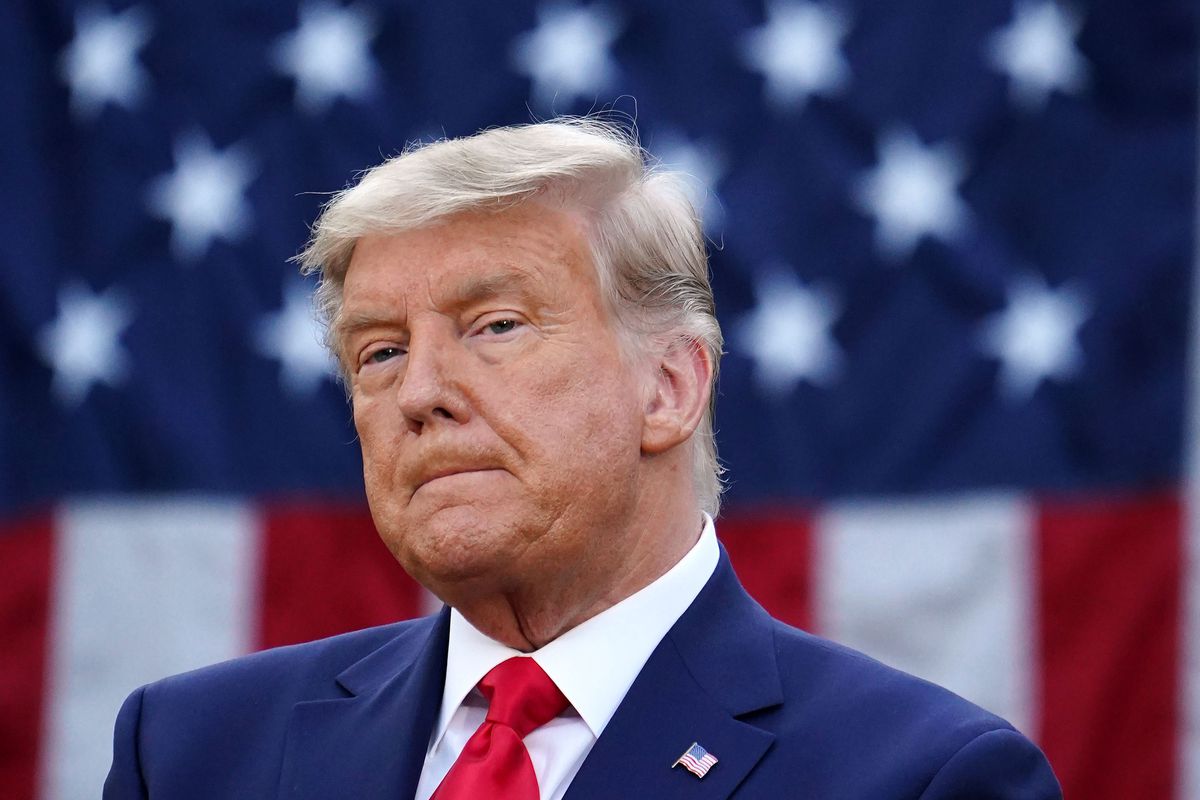 Trump, in a navy suit, white shirt, and red tie, frowns slightly as he sucks his lips. His hair looks more silver than usual, and he is in front of a US flag.