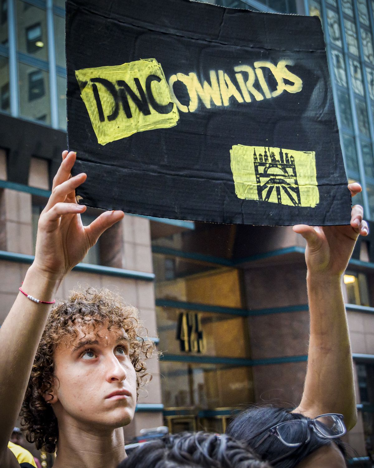 A young person holds a sign over their head that reads, “DNCowards.”