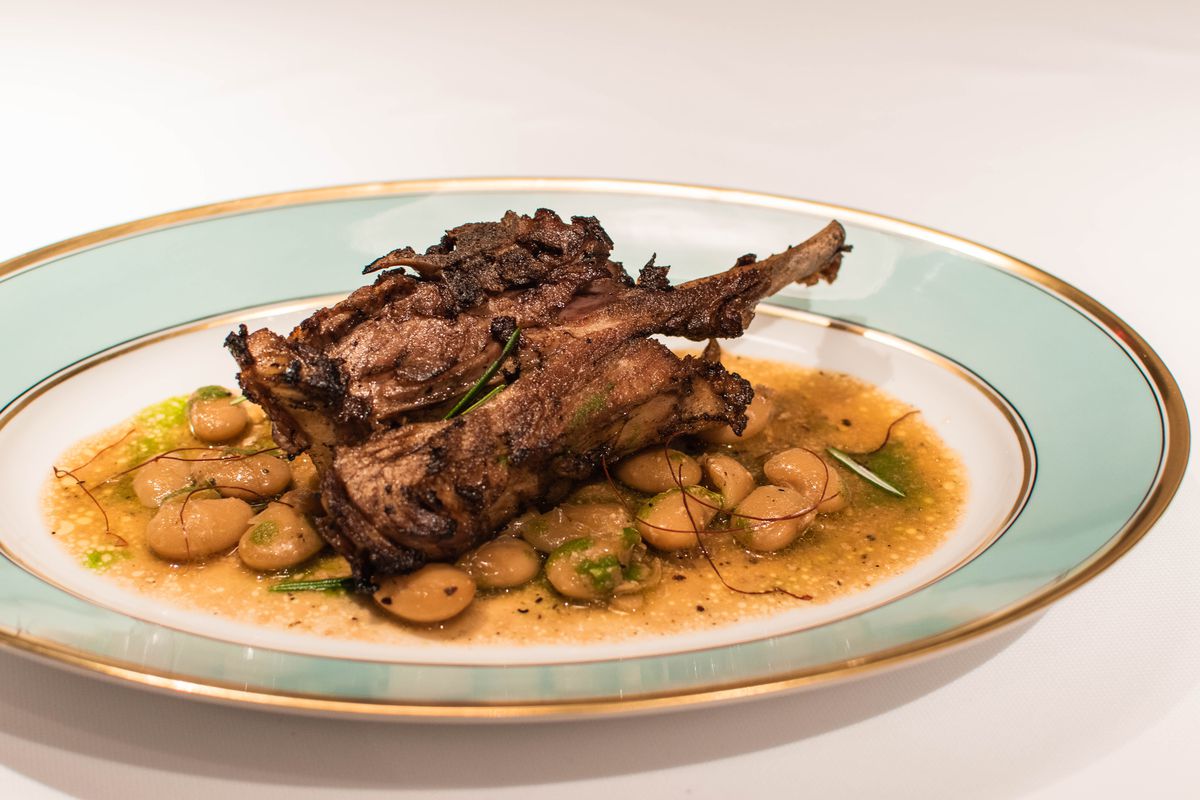 A slow-cooked piece of lamb sits in an oval plate with a gold and green trim. It sits on a bed of beans bathed in a yellow sauce.