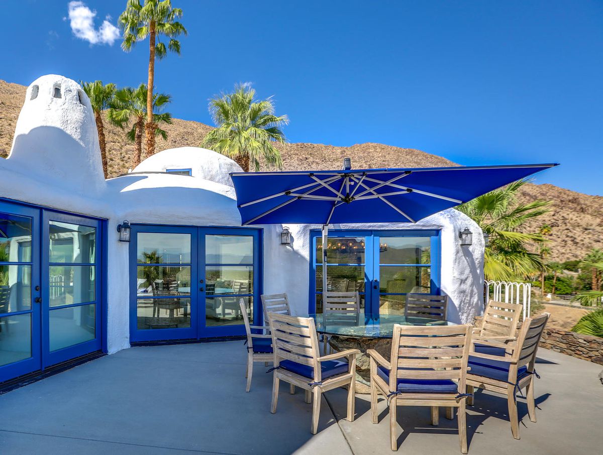 An outdoor dining area has teak chairs with blue cushions, a blue umbrella, and a glass table top. 