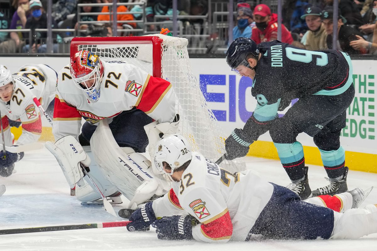 Ryan Donato, on the right hand of the frame, is behind the net and trying to sneak the puck past Sergei Bobrovsky. A Panthers player is sprawled out on the ice also trying to stop the play. Donato is in the Kraken’s home navy jersey and the Panthers’ players are in away white jerseys.