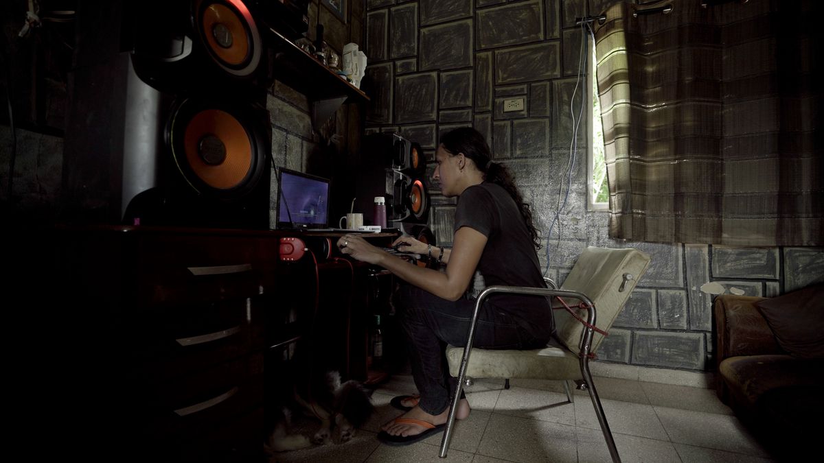 A young man with tied back black hair sits in a white chair in a dimly lit stonewalled room in Cuba playing a video game on a small laptop computer