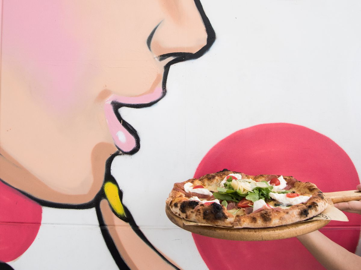 A pizza is held up to a wall mural depicting a woman. It looks like she’s about to eat the pizza.