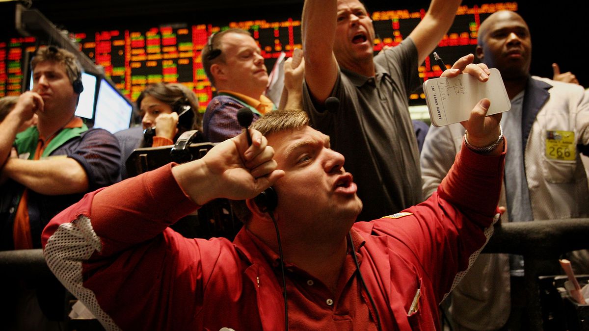A swarm of male option traders shout and gesture, most with hands in the air signaling trades, against a backdrop of large monitor screens showing strings of color-coded prices.