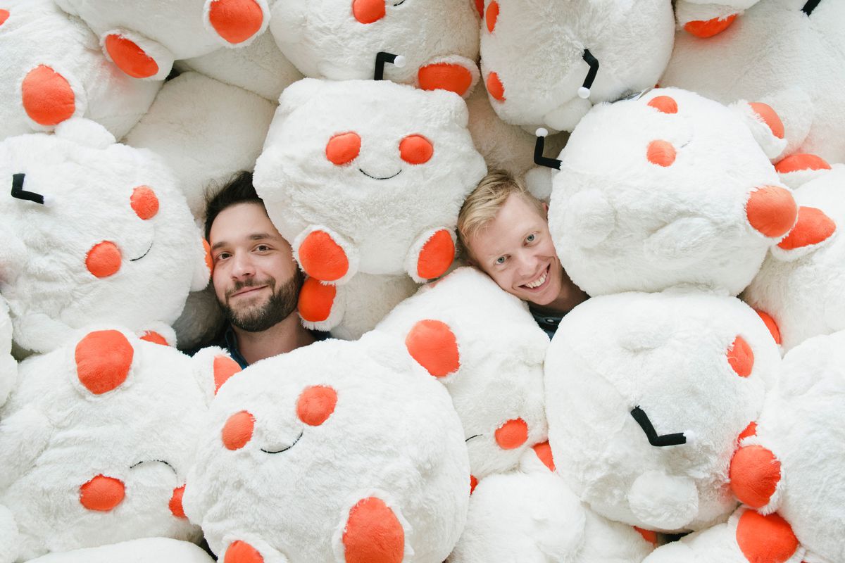 A caucasian man with blonde hair smiles out from a pile of plush toys shaped like the Reddit logo.