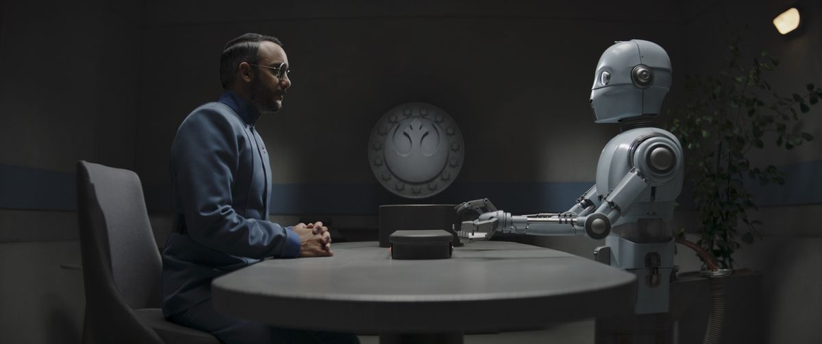 El Dr. Pershing se sienta frente a un robot blanco y delgado en una mesa gris en The Mandalorian
