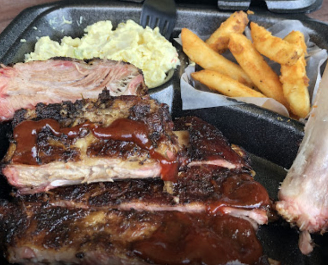 A black takeout plate holds ribs drizzled with barbecue sauce and sides of fries and potato salad.