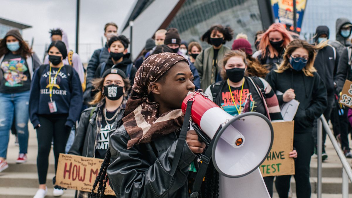 A young woman wearing a black jacket and brown headdress speaks through a red and white bullhorn microphone. There are several students holding signs or standing in solidarity behind her.