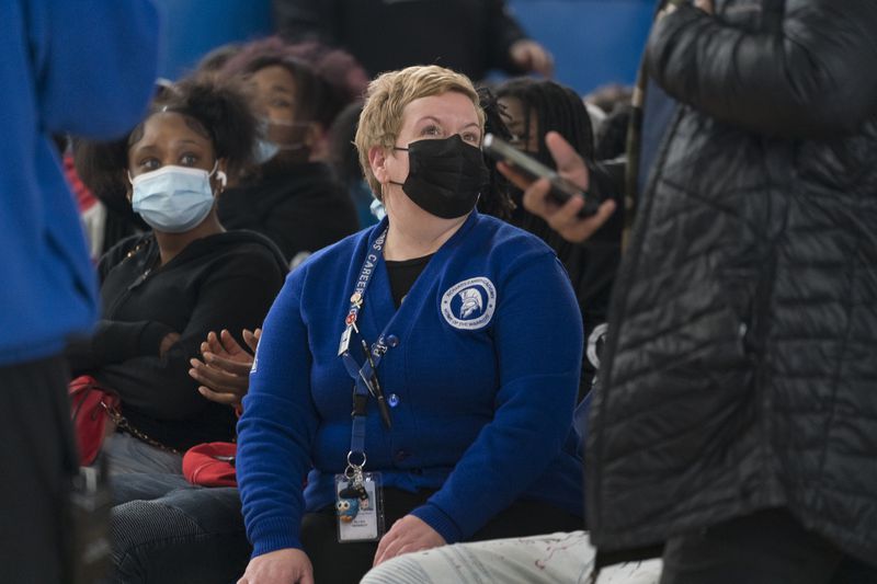 A school principal, wearing a blue Richards High School sweater and black mask, looks over at a person in the foreground while sitting in a chair amongst her students.