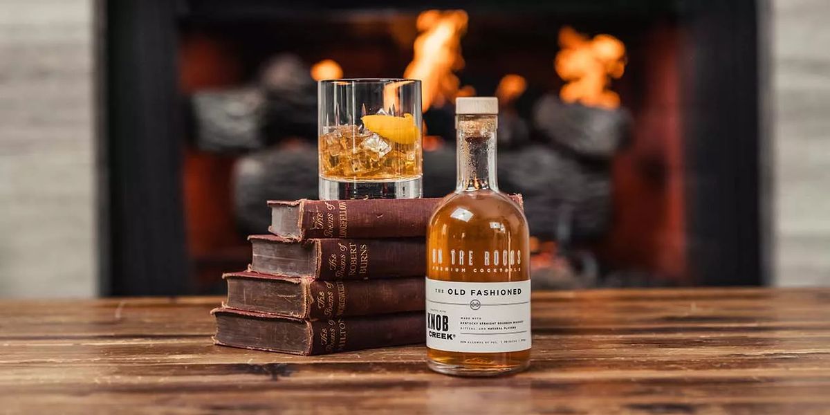A pre-mixed Old Fashioned cocktail is in a bottle, in front of a fireplace and next to a stack of books.