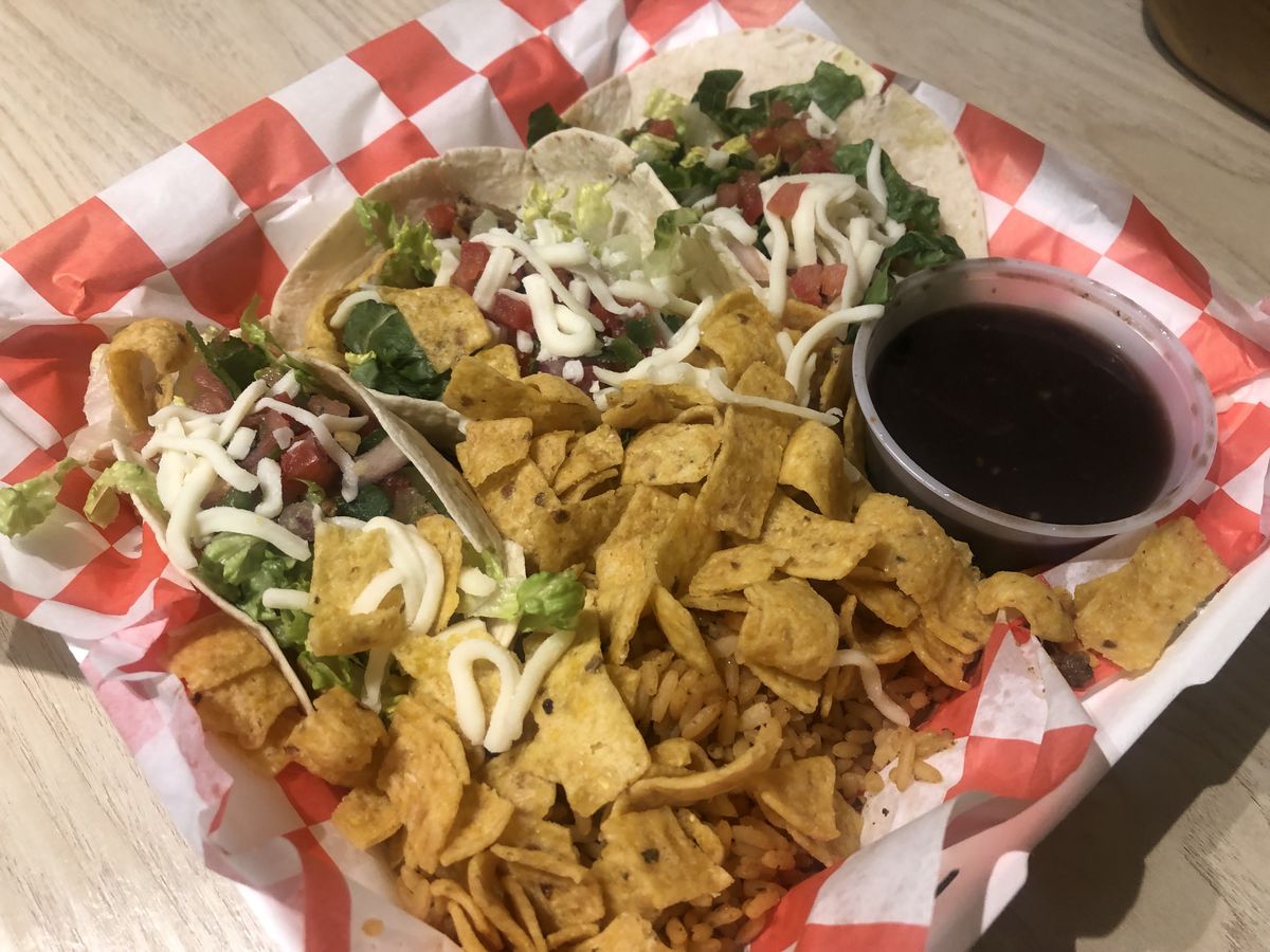 Three tacos, a cup of black beans, and a scoop of Fritos arranged in a styrofoam takeout container lined in red and white checkered paper.