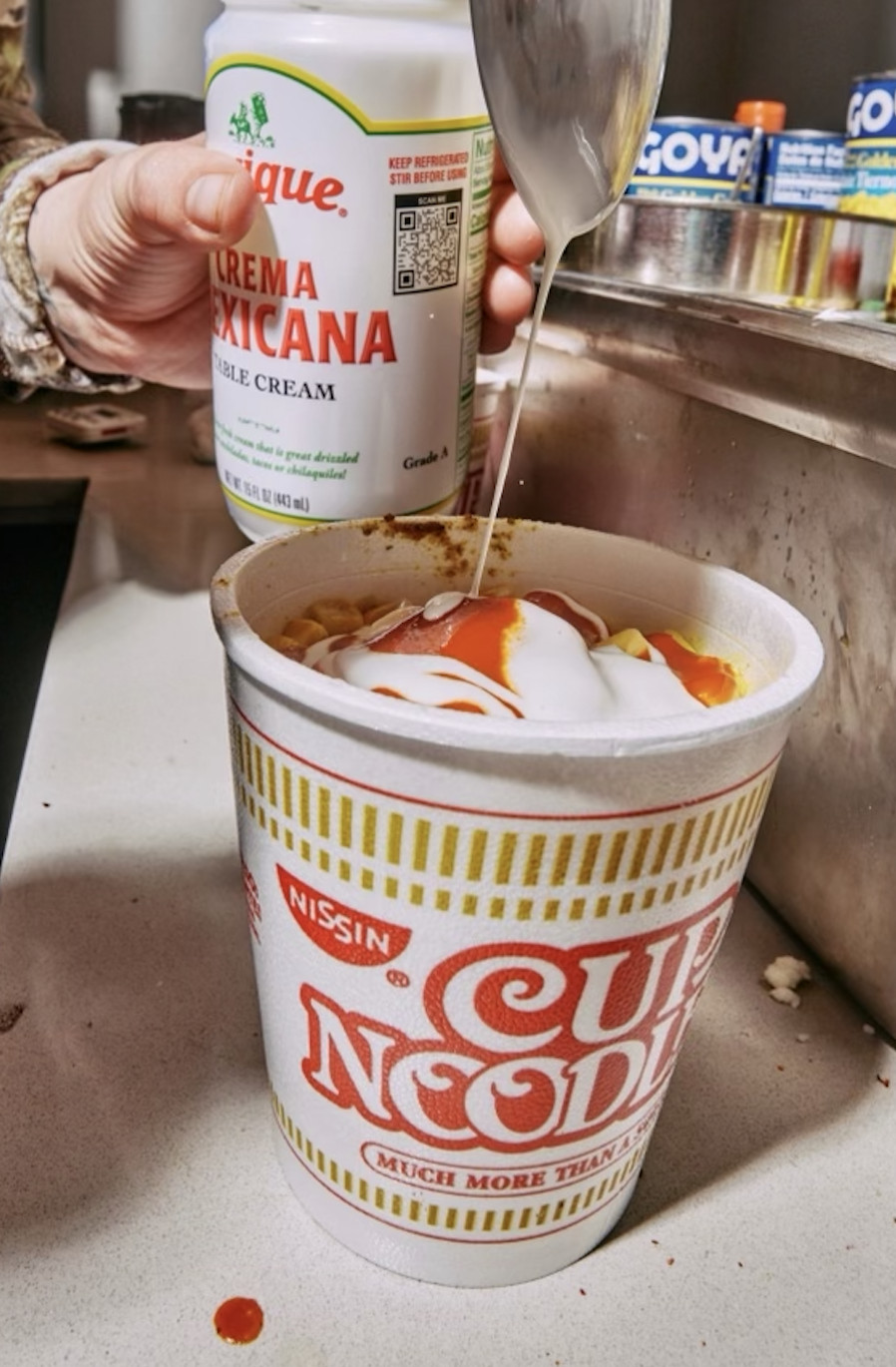 A hand pours crema Mexicana into a Cup Noodles. 