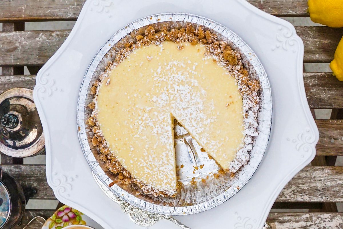 A pie in a metal tin with a cookie crust and a creamy yellow filling with one slice removed. It’s dusted with powdered sugar. 