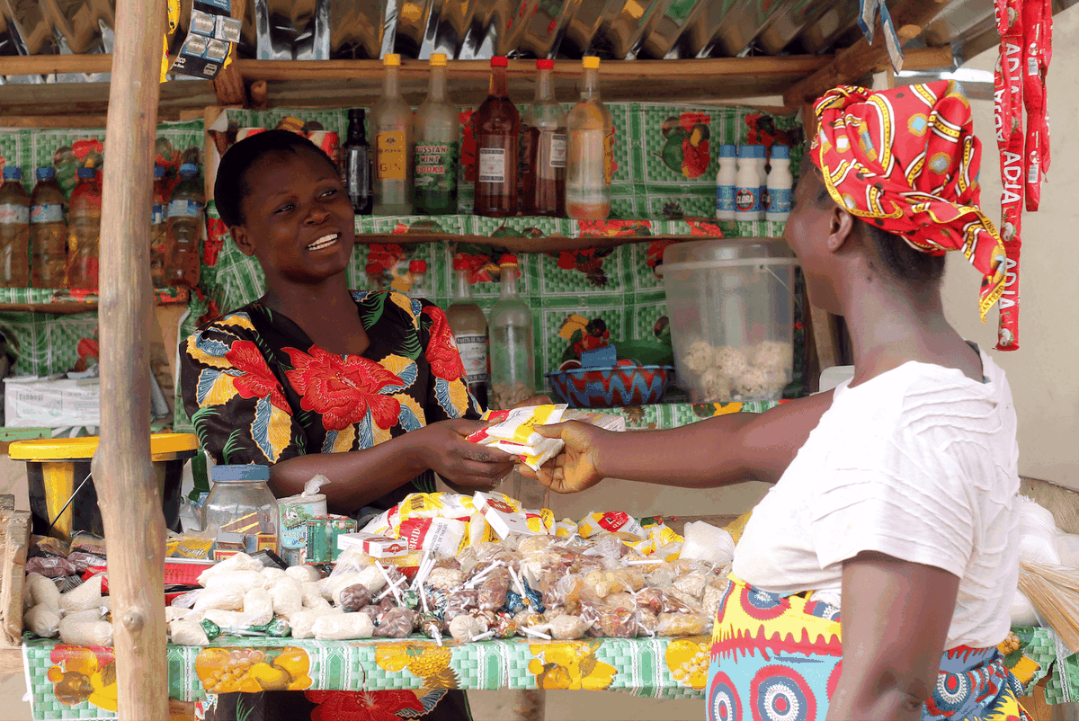 Two women transact a sale across a counter covered in goods.