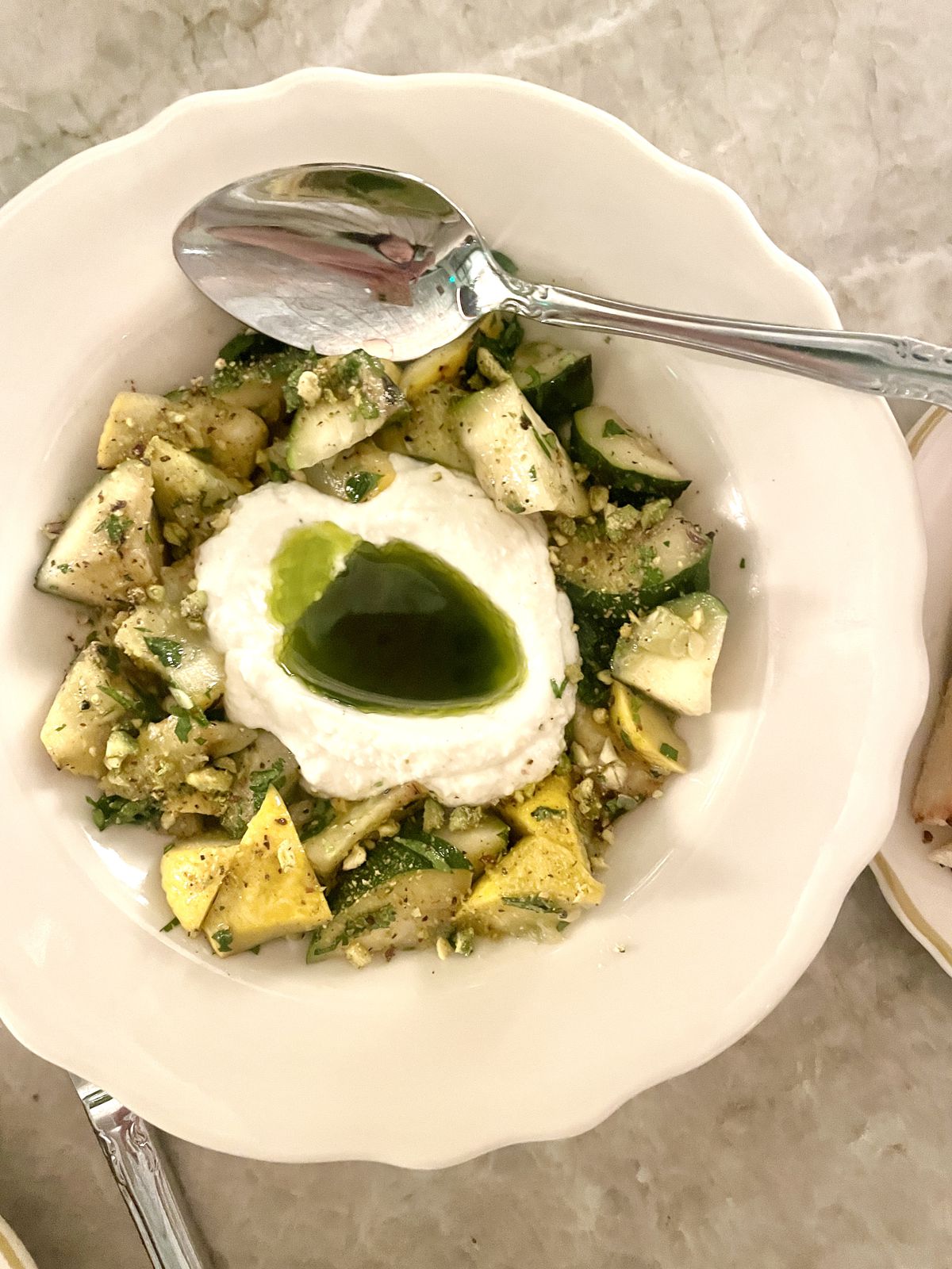 A bowl holds ricotta in the center with a green-infused oil. Around it sits grilled zucchini. A silver spoon is dipped in on one side.
