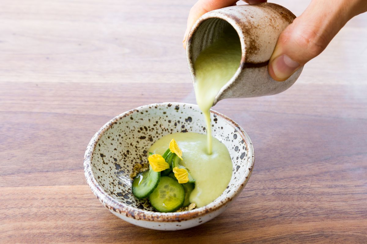 Green soup being poured into ceramic bowl over sliced cucumbers.