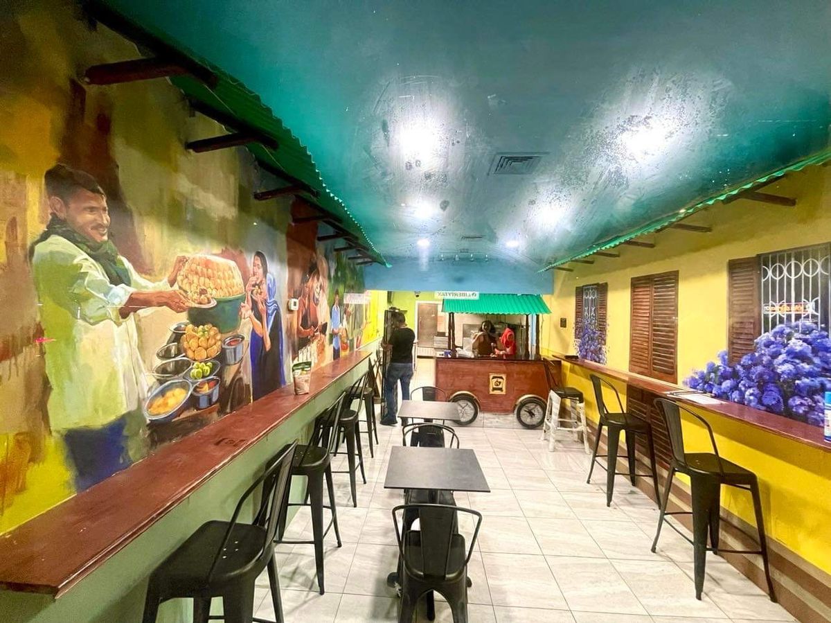 The interior of a restaurant shows stools along the left and right mural-painted walls, tables down the center, and a street cart illustration in the back.