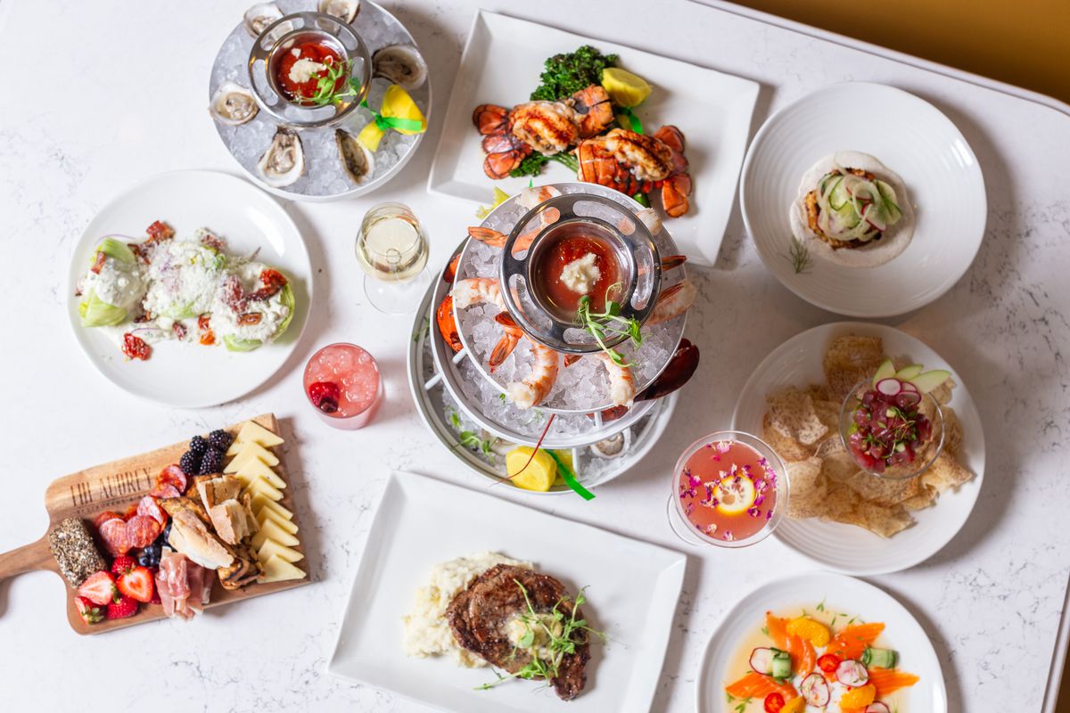 A table at the Audrey Restaurant &amp; Bar with a seafood tower, a wedge salad topped with parmesan, a platter of oysters on the half shell, lobster tails, a steak over potatoes, a charcuterie board, and more.