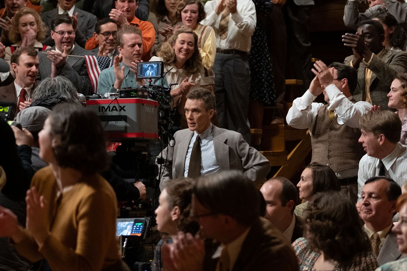 A photo of Cillian Murphy in a crowd acting in front of an IMAX camera.