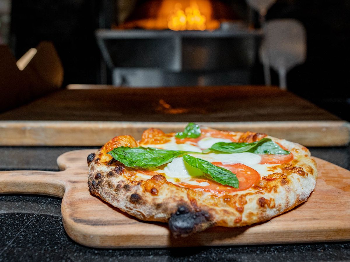 A small puffy Margherita pizza on a wooden peel in front of a wood-fired oven.