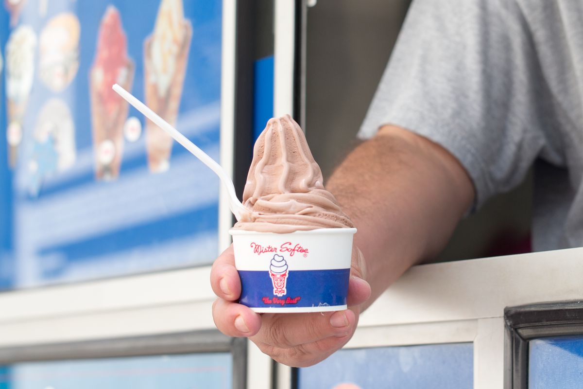 A man holds a cup of chocolate soft serve in a Mister Softee cup with a spoon in it.
