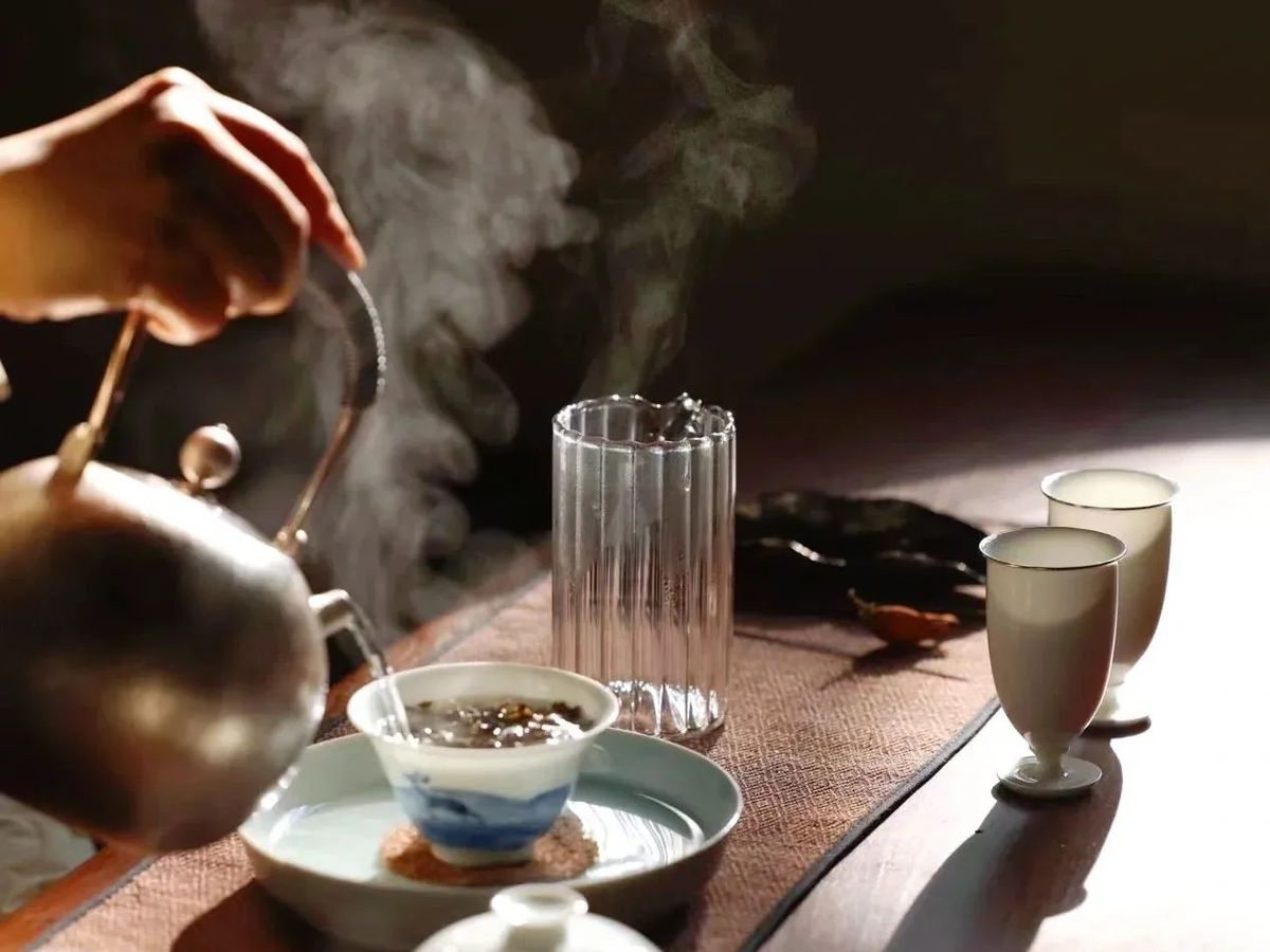 A hand pours tea from a kettle into a delicate cup in a room with dim lighting. A lot of steam rises from the cup.