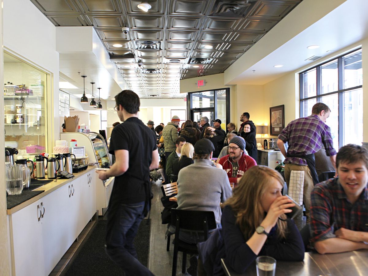 A restaurant’s interior with white walls, a counter to the left, and diners seated at tables to the right. 