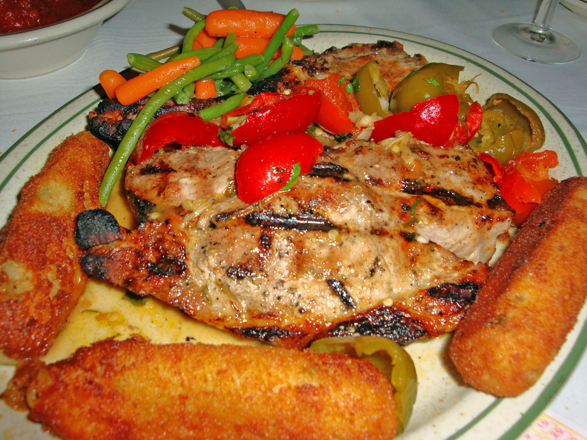 A white and green dish with pork chops in the middle surrounded by oblong croquettes and a side serving of tomatoes, green beans, and carrots