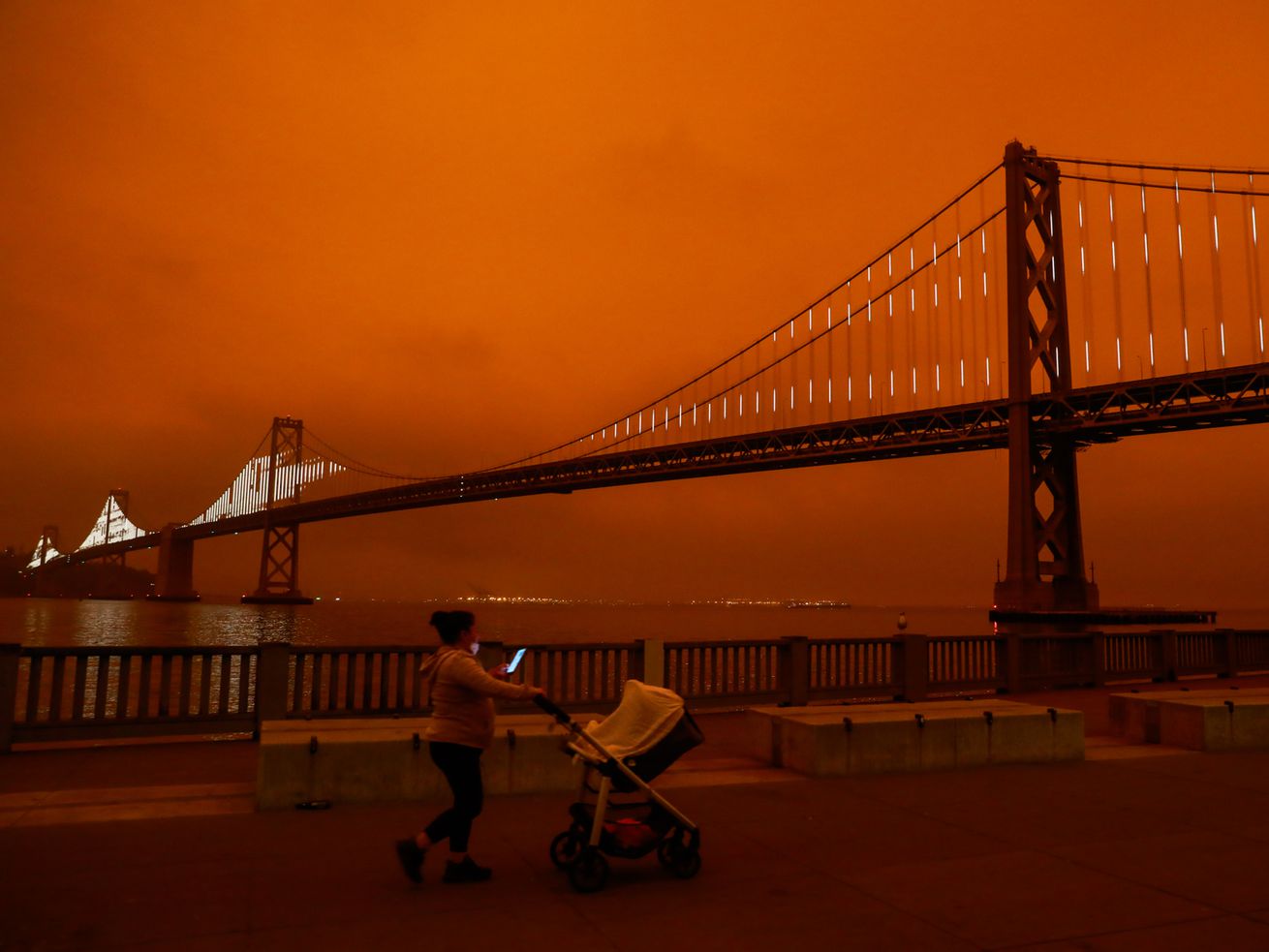 A person pushes a stroller along the waterfront in San Francisco with the Bay Bridge and an orange sky behind them.