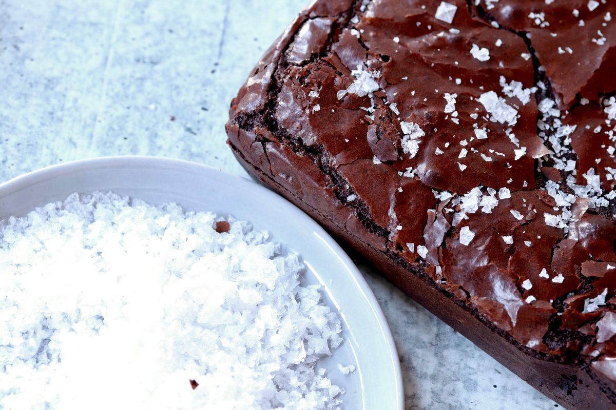 A close-up picture of a bowl of sea salt next to a big block of brownie with sea salt on top.