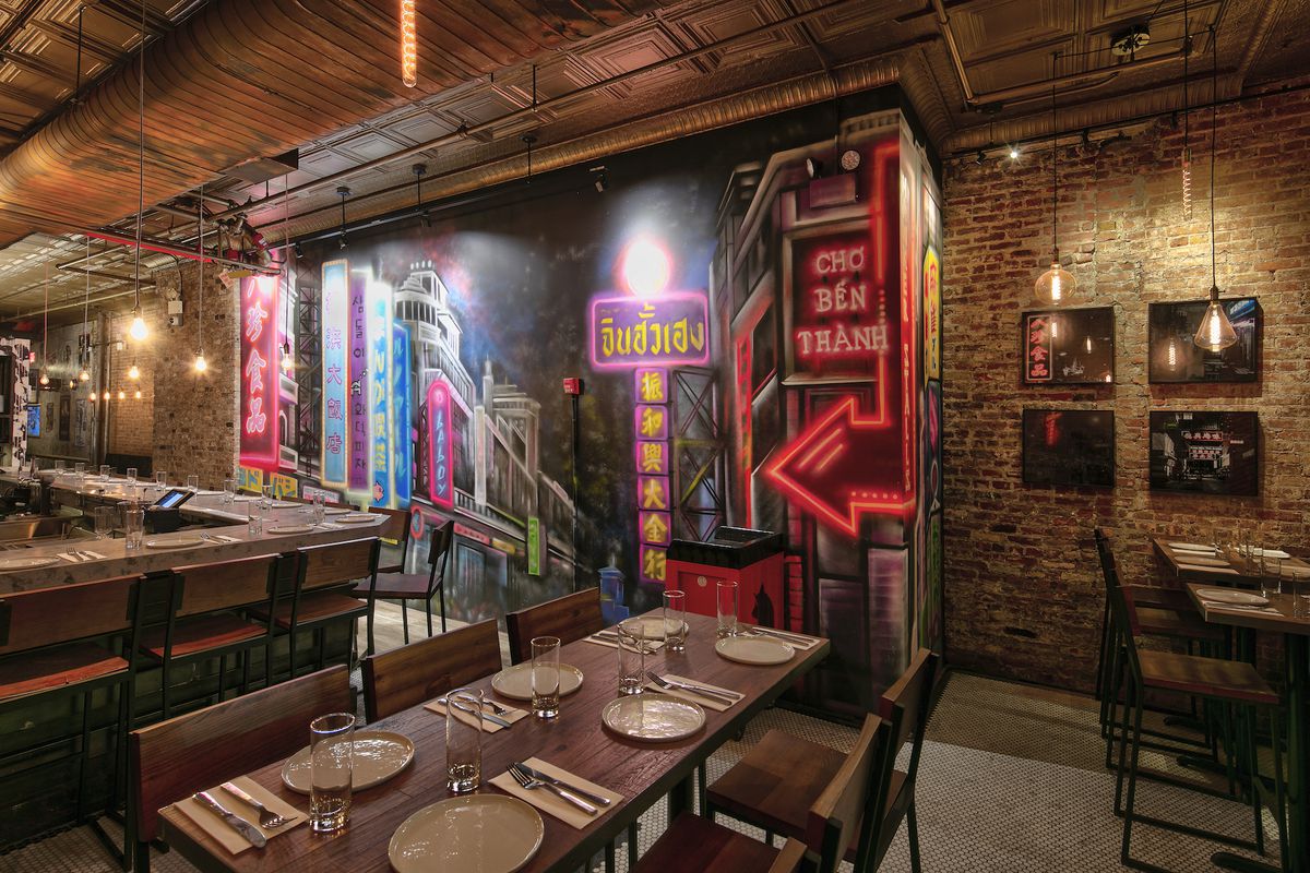 The interior of a restaurant with exposed brick walls, wooden tables placed closed together, and a mural depicting street food markets in Asia