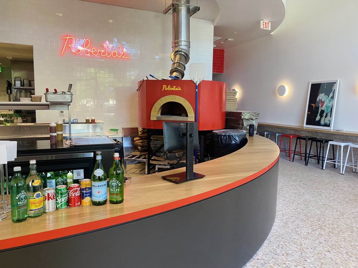 A counter lined with sodas, in front of a red wood-fired pizza oven.