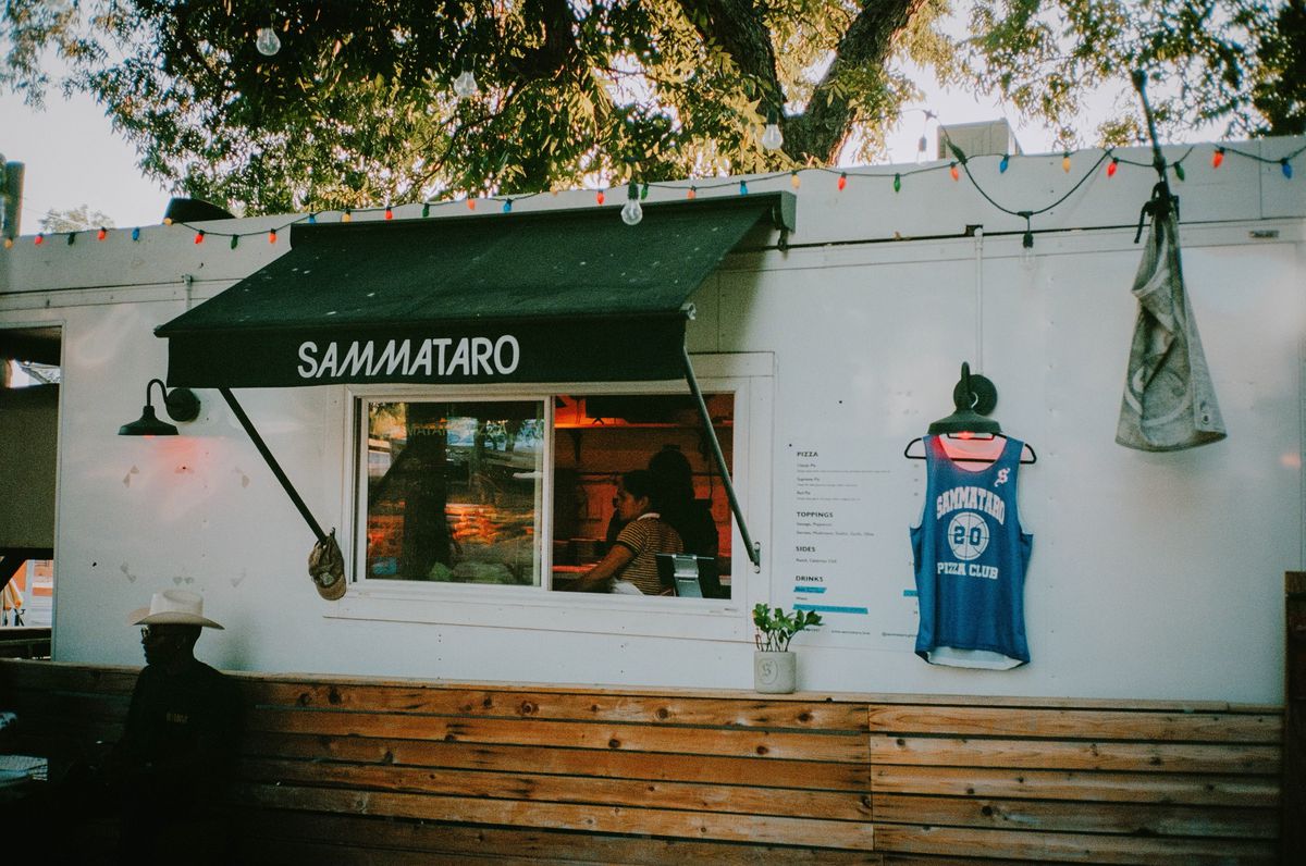 A white food truck with a window and a black awning reading “Sammataro” and a hanging blue basketball jersey.