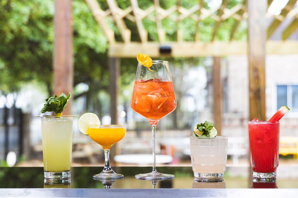 Multiple colorful cocktails lines in a row on a bar top with a patio scenery slightly out of focus in the background.