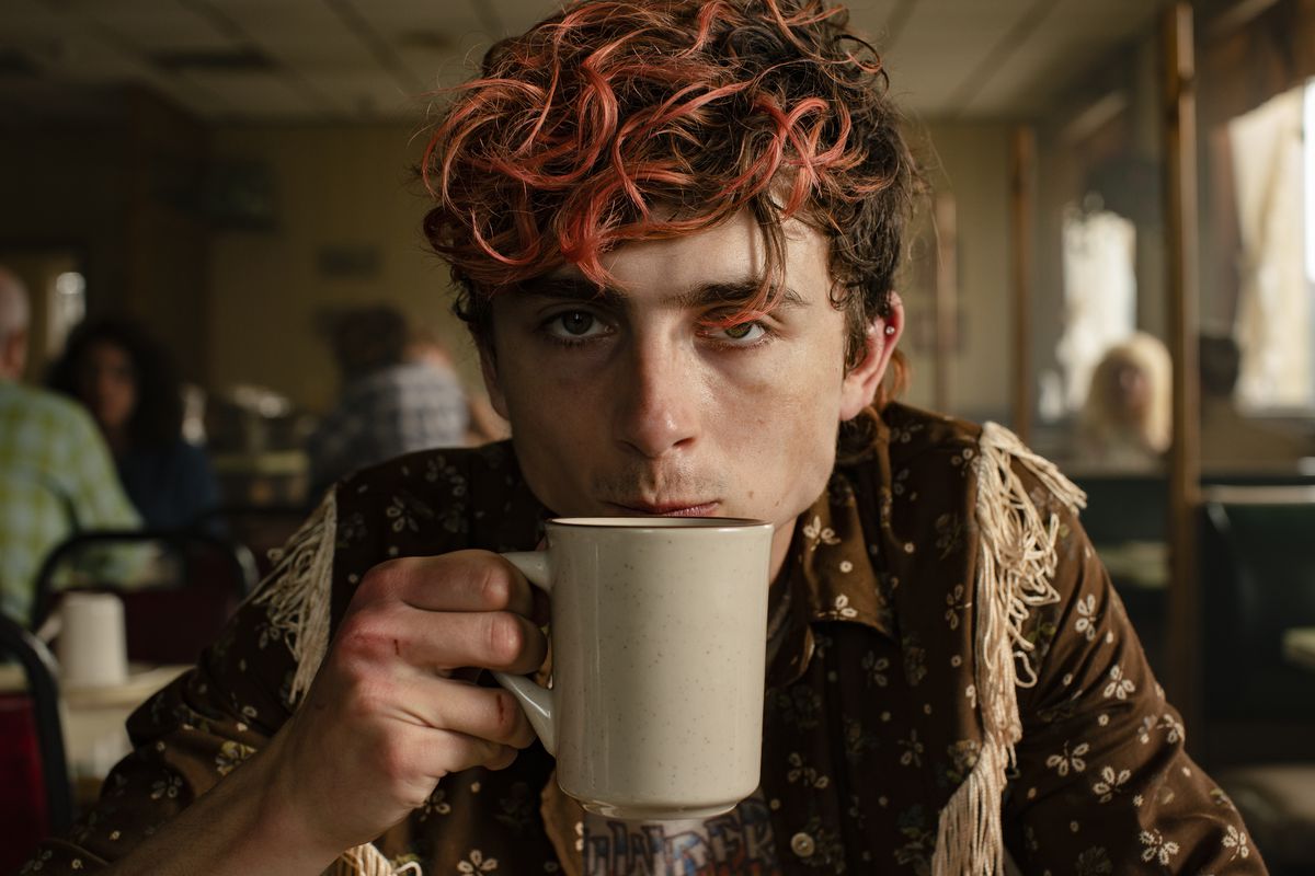 Lee (Timothée Chalamet), un joven con profundas bolsas en los ojos y una mata de pelo rizado teñido de rojo, bebe café y mira a la cámara con cara de confrontación en Bones and All