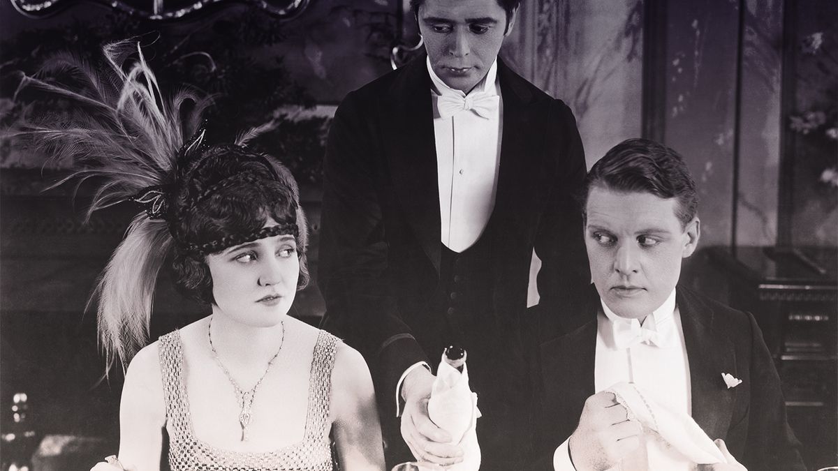 A man and a woman dressed up for a 1920s dinner party give each other the side eye as a waiter reaches between them awkwardly to refill their wine.