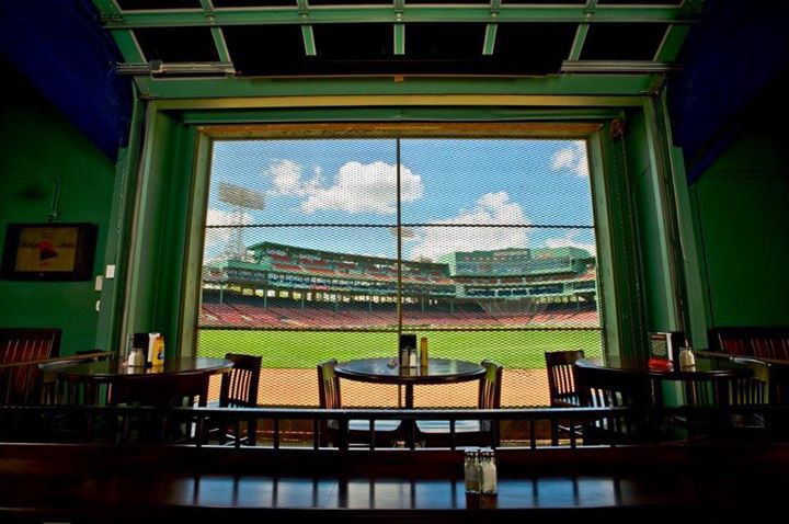 Interior of a bar with green walls. A large window looks out onto an empty baseball stadium on a sunny day.