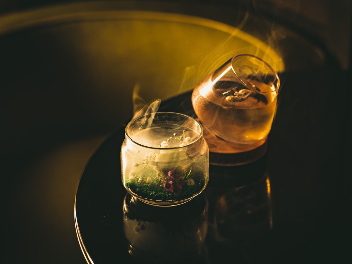 Two rocks glasses, one with smoke and one with an Old Fashioned set on a dark tabletop with moody lighting.